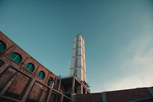 A towering skyscraper reaches into a clear blue sky, surrounded by adjacent buildings with large arched windows. The scene conveys a sense of architectural progress and urban development.