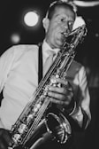 A person wearing a light-colored shirt plays a saxophone, captured in a close-up black and white photograph. The musician's expression is focused, and the instrument's gleaming keys and details are highlighted by the lighting.