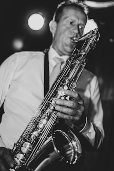 A person wearing a light-colored shirt plays a saxophone, captured in a close-up black and white photograph. The musician's expression is focused, and the instrument's gleaming keys and details are highlighted by the lighting.