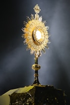 A golden, ornate religious object with a sunburst design, positioned on a dark background. The central section glows with a bright, circular light surrounded by intricate detailing and gems.