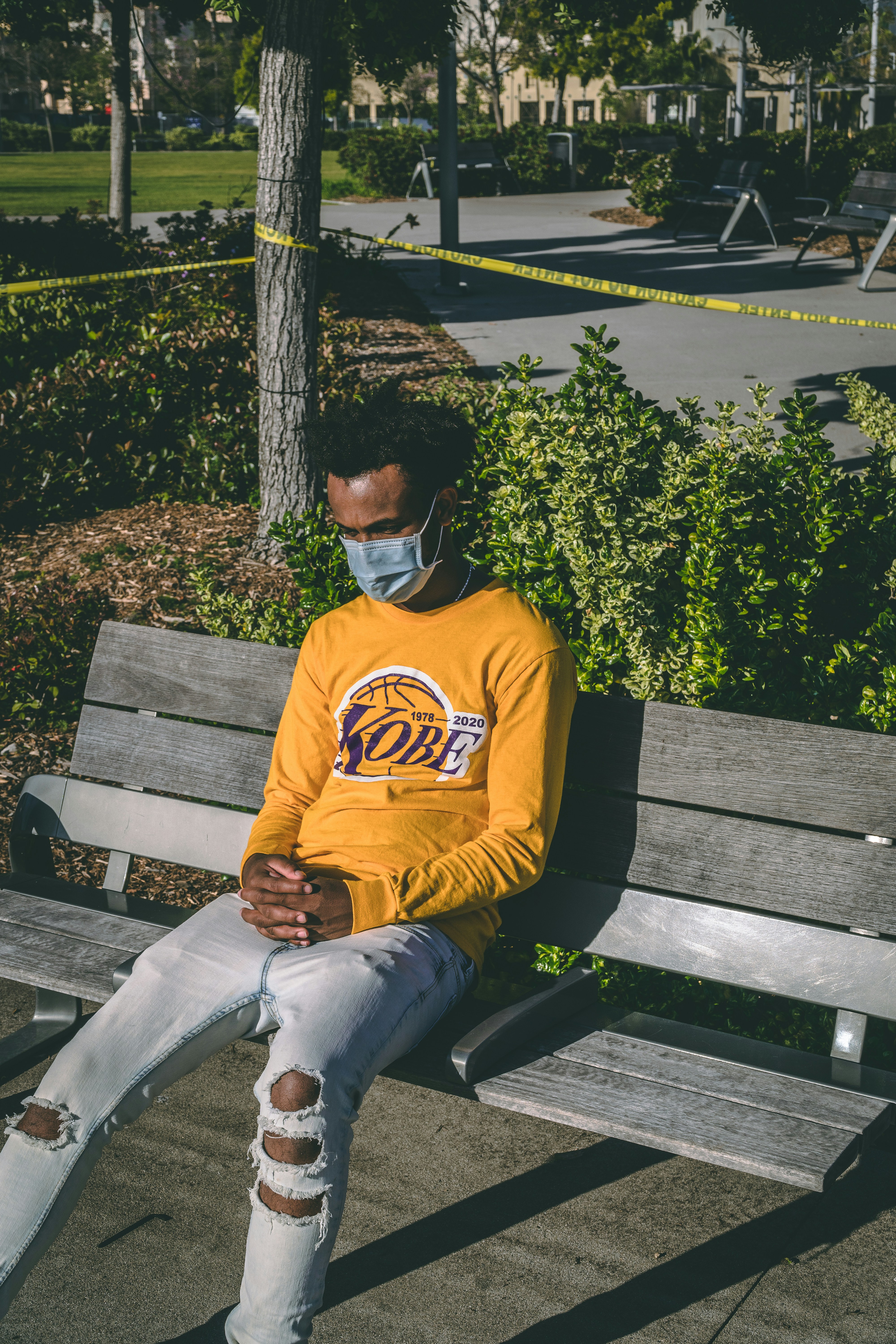 A man wearing a Lakers shirt and ripped jeans and a face mask sitting on a bench.