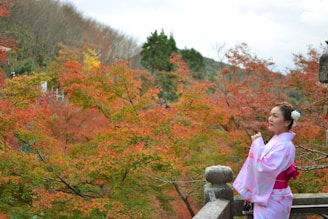A woman dressed in a light pink kimono with floral patterns stands on a stone balcony against a backdrop of vibrant autumn foliage. The leaves exhibit a range of colors from green to deep red. In the background, a dense cluster of trees covers rolling hills under a partially cloudy sky.