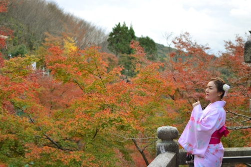 A woman dressed in a light pink kimono with floral patterns stands on a stone balcony against a backdrop of vibrant autumn foliage. The leaves exhibit a range of colors from green to deep red. In the background, a dense cluster of trees covers rolling hills under a partially cloudy sky.