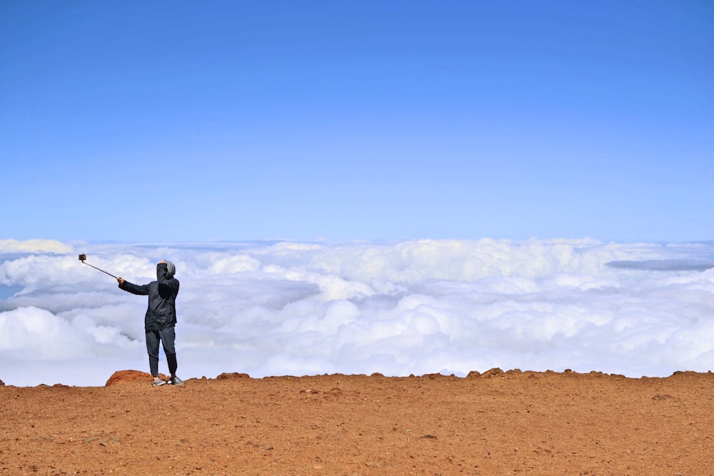 man in black jacket and pants standing on brown field under white clouds and blue sky