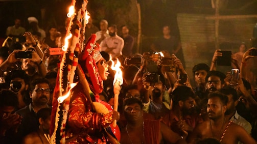 A vibrant scene depicting a traditional cultural event with a performer in ornate attire holding torches. The performer is surrounded by a large crowd of people, many of whom are capturing the event on their phones. The setting appears festive and lively, with warm lighting enhancing the atmosphere.