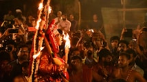 A vibrant scene depicting a traditional cultural event with a performer in ornate attire holding torches. The performer is surrounded by a large crowd of people, many of whom are capturing the event on their phones. The setting appears festive and lively, with warm lighting enhancing the atmosphere.