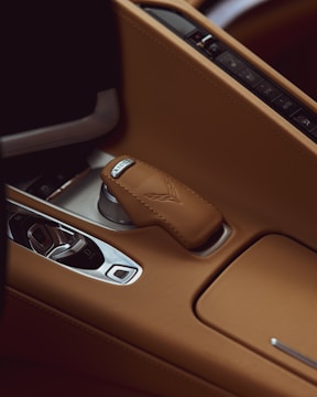 A close-up of a car's interior highlighting the gear selector encased in tan leather with distinctive stitching. The surrounding console is also covered with the same tan leather. A metallic knob and several buttons are visible, contributing to a luxurious automotive design.