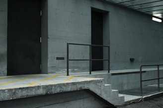 The image depicts a desolate urban setting with concrete structures. A loading dock area features a ramp, steps, and a metal railing. The walls are grey, and two large doors are visible, one of which is slightly ajar. A fluorescent light is mounted on the wall, casting a dim glow over the scene.