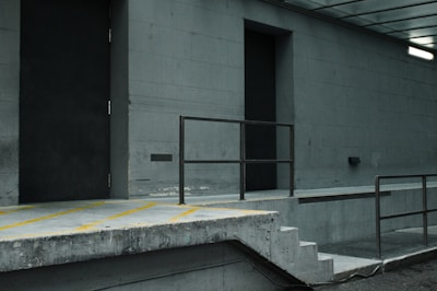 The image depicts a desolate urban setting with concrete structures. A loading dock area features a ramp, steps, and a metal railing. The walls are grey, and two large doors are visible, one of which is slightly ajar. A fluorescent light is mounted on the wall, casting a dim glow over the scene.