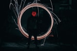 A mysterious figure stands in a dimly lit environment, wearing a mask with glowing red patterns resembling a face with crossed eyes and a smile. Bright, swirling light trails form a circle around the person, creating a captivating effect against the dark background.