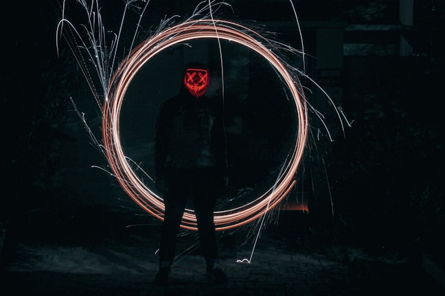 A mysterious figure stands in a dimly lit environment, wearing a mask with glowing red patterns resembling a face with crossed eyes and a smile. Bright, swirling light trails form a circle around the person, creating a captivating effect against the dark background.