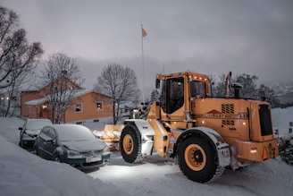 A snow-covered landscape featuring a large orange snowplow clearing the road next to parked cars, one of which is also covered in snow. Nearby, a warmly-lit house sits surrounded by bare trees, with overcast skies contributing to a cold, wintry atmosphere.
