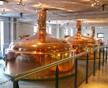 Large, polished copper brewing tanks in an indoor brewery setting with overhead lighting and a railing in the foreground. The room has white walls and several windows allowing natural light to complement the artificial lighting.