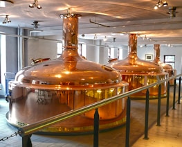 Large, polished copper brewing tanks in an indoor brewery setting with overhead lighting and a railing in the foreground. The room has white walls and several windows allowing natural light to complement the artificial lighting.
