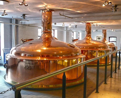 Large, polished copper brewing tanks in an indoor brewery setting with overhead lighting and a railing in the foreground. The room has white walls and several windows allowing natural light to complement the artificial lighting.