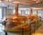 Large, polished copper brewing tanks in an indoor brewery setting with overhead lighting and a railing in the foreground. The room has white walls and several windows allowing natural light to complement the artificial lighting.