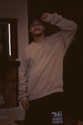 A person stands indoors wearing a casual beige sweatshirt and black shorts, with a cap and light facial hair. The lighting is dim and warm, creating a cozy atmosphere. The background is mostly dark, with some indistinct features.