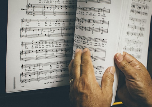Hands are holding and flipping through a page of sheet music, featuring musical notes and lyrics. The page is slightly worn and situated next to a piano.