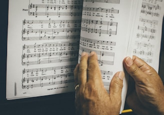 Hands are holding and flipping through a page of sheet music, featuring musical notes and lyrics. The page is slightly worn and situated next to a piano.