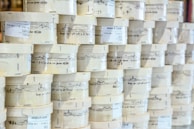 Rows of cylindrical wooden boxes are stacked, each labeled with printed text and graphics. The boxes appear to contain cheese, indicated by the words and imagery on the packaging.