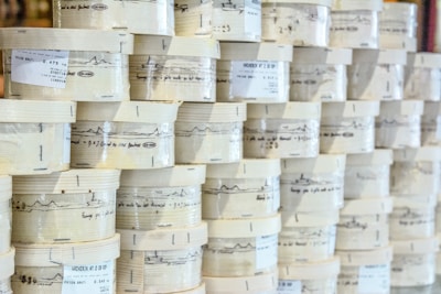 Rows of cylindrical wooden boxes are stacked, each labeled with printed text and graphics. The boxes appear to contain cheese, indicated by the words and imagery on the packaging.