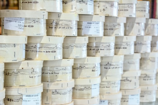 Rows of cylindrical wooden boxes are stacked, each labeled with printed text and graphics. The boxes appear to contain cheese, indicated by the words and imagery on the packaging.