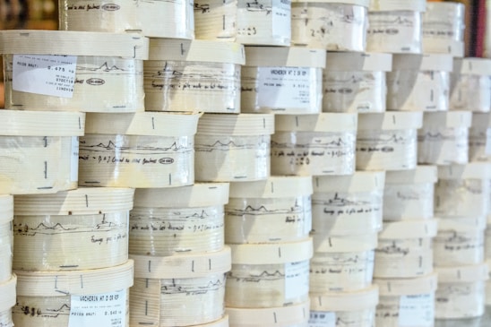 Rows of cylindrical wooden boxes are stacked, each labeled with printed text and graphics. The boxes appear to contain cheese, indicated by the words and imagery on the packaging.