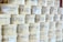 Rows of cylindrical wooden boxes are stacked, each labeled with printed text and graphics. The boxes appear to contain cheese, indicated by the words and imagery on the packaging.
