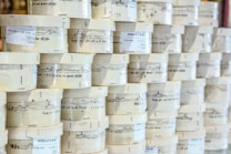 Rows of cylindrical wooden boxes are stacked, each labeled with printed text and graphics. The boxes appear to contain cheese, indicated by the words and imagery on the packaging.