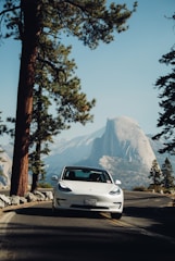 A white Tesla car is parked on a road surrounded by tall pine trees. In the background, a large granite mountain rises under a clear blue sky, suggesting a scenic location such as a national park.