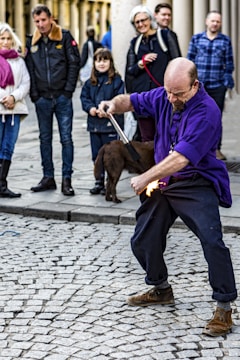 A street performer dressed in a purple shirt skillfully manipulates a stick with flames at one end, captivating an audience of onlookers. The crowd, including men, women, and a child, watch with interest. The scene takes place on a cobbled pedestrian street, with some people accompanied by a dog.