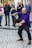 A street performer dressed in a purple shirt skillfully manipulates a stick with flames at one end, captivating an audience of onlookers. The crowd, including men, women, and a child, watch with interest. The scene takes place on a cobbled pedestrian street, with some people accompanied by a dog.