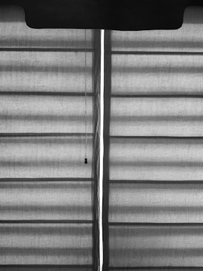Horizontal blinds with a fabric texture are partially drawn, allowing a vertical sliver of light to pass through. A pull cord hangs from the upper portion of the blinds.