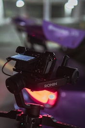 A camera is mounted on a gimbal with the brand name Ronin visible. The background is blurred, with bright circular lights and a purple object, possibly a car, in the background. The environment seems dimly lit with a soft focus on the camera and gimbal.