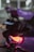 A camera is mounted on a gimbal with the brand name Ronin visible. The background is blurred, with bright circular lights and a purple object, possibly a car, in the background. The environment seems dimly lit with a soft focus on the camera and gimbal.