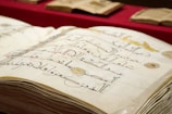 A close-up of an open book displaying Arabic calligraphy on yellowed pages. The writing is ornate and decorative, with some colorful accents. Several other similar-looking books are in the background, placed on a red surface.