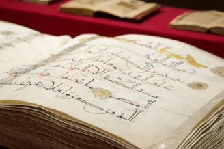 A close-up of an open book displaying Arabic calligraphy on yellowed pages. The writing is ornate and decorative, with some colorful accents. Several other similar-looking books are in the background, placed on a red surface.