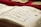 A close-up of an open book displaying Arabic calligraphy on yellowed pages. The writing is ornate and decorative, with some colorful accents. Several other similar-looking books are in the background, placed on a red surface.