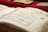 A close-up of an open book displaying Arabic calligraphy on yellowed pages. The writing is ornate and decorative, with some colorful accents. Several other similar-looking books are in the background, placed on a red surface.