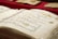 A close-up of an open book displaying Arabic calligraphy on yellowed pages. The writing is ornate and decorative, with some colorful accents. Several other similar-looking books are in the background, placed on a red surface.