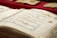 A close-up of an open book displaying Arabic calligraphy on yellowed pages. The writing is ornate and decorative, with some colorful accents. Several other similar-looking books are in the background, placed on a red surface.
