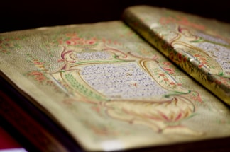 An open book with intricately detailed, decorative pages featuring elaborate floral and ornate designs along with written script. The artwork includes elements in gold and is complemented by vivid colors such as red, green, and blue.