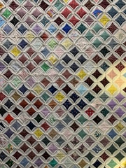 A quilt composed of numerous small, colorful fabric squares connected by a white lattice pattern. Each square features a unique design, ranging from polka dots and floral patterns to checkered designs. The quilt combines various colors including reds, greens, blues, and purples to create a vibrant patchwork effect.