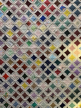 A quilt composed of numerous small, colorful fabric squares connected by a white lattice pattern. Each square features a unique design, ranging from polka dots and floral patterns to checkered designs. The quilt combines various colors including reds, greens, blues, and purples to create a vibrant patchwork effect.