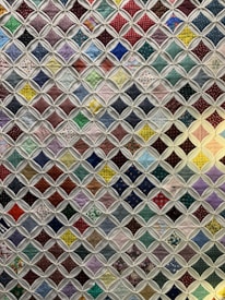 A quilt composed of numerous small, colorful fabric squares connected by a white lattice pattern. Each square features a unique design, ranging from polka dots and floral patterns to checkered designs. The quilt combines various colors including reds, greens, blues, and purples to create a vibrant patchwork effect.