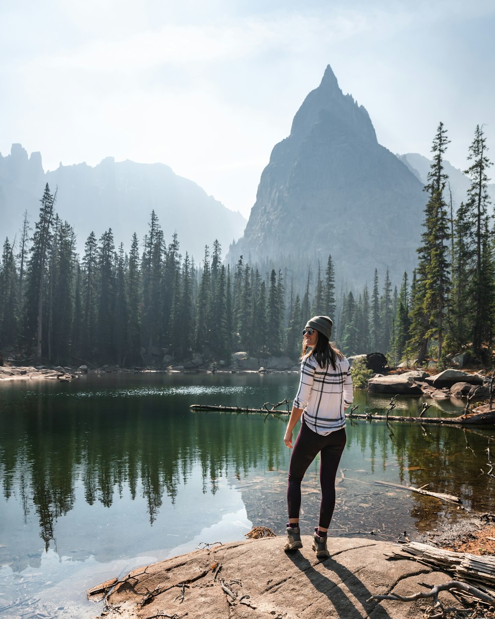 woman in white and pink plaid shirt and black pants standing on rock near lake during