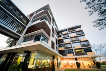 A modern multi-story office building with a sleek, architectural design. The facade features a combination of glass and horizontal wooden slats, providing a contemporary aesthetic. Large windows and prominent balconies enhance the structure's open and airy feel. The image is taken from a low angle, emphasizing the building's height and geometric lines. The surroundings include landscaped greenery with a couple of trees visible.