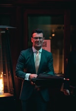 A man in a suit and tie is standing indoors holding an umbrella. He has glasses and is positioned near a tall heat lamp with a visible flame. The environment is dimly lit, creating a warm, intimate atmosphere with dark tones and soft light.