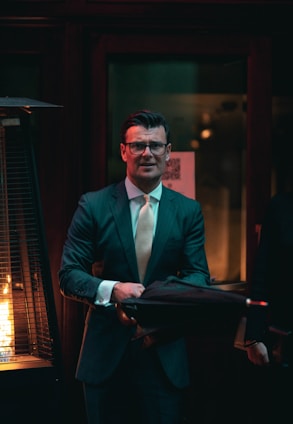 A man in a suit and tie is standing indoors holding an umbrella. He has glasses and is positioned near a tall heat lamp with a visible flame. The environment is dimly lit, creating a warm, intimate atmosphere with dark tones and soft light.
