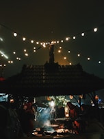 A cozy outdoor setting with a group of people gathered around a fire pit under a wooden shelter. String lights hang above, illuminating the scene with a warm glow. Smoke rises from the fire, adding to the ambiance. In the background, there are festive decorations and greenery, suggesting a holiday or winter market setting.